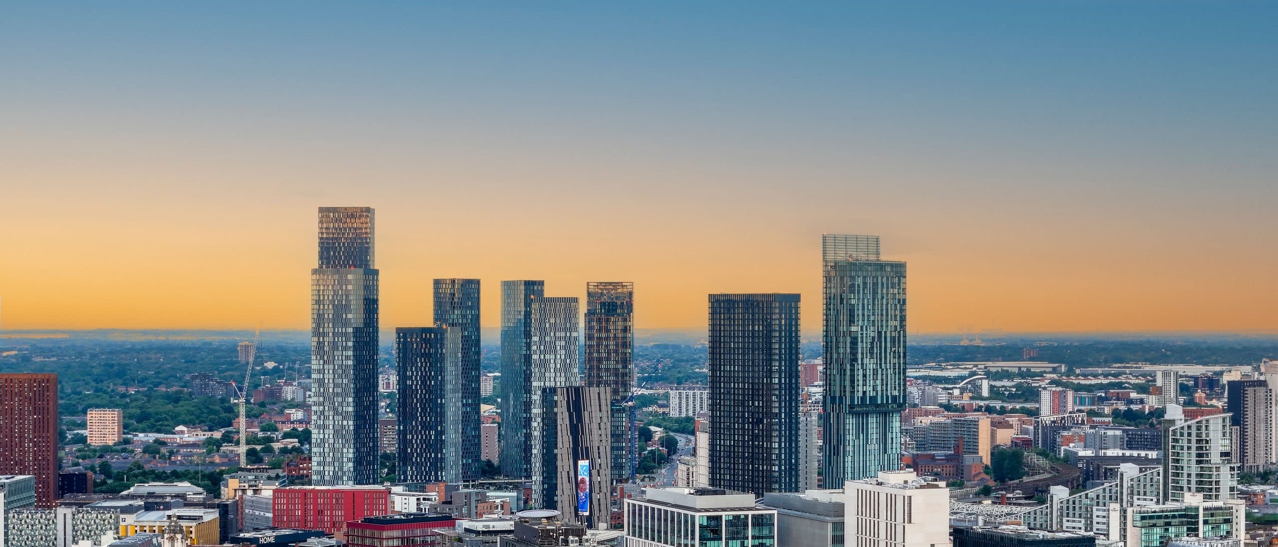 Towers of Manchester city centre standing against a clear sky that fades from blue to orange at the horizon, with open sky above for text.