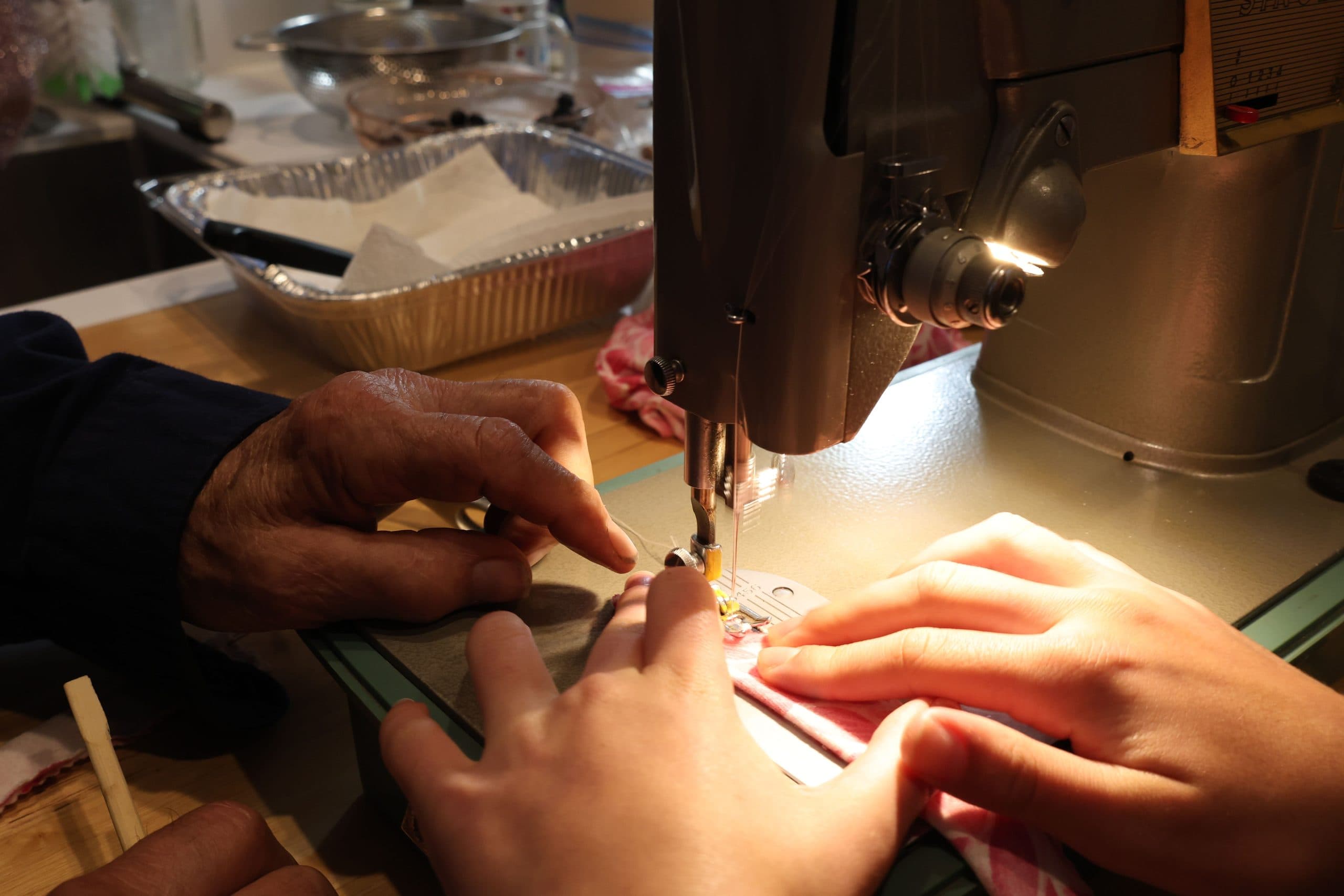 Close view of an older hand and a younger hand guiding fabric under the needle of a vintage sewing machine, lit by a warm lamp.