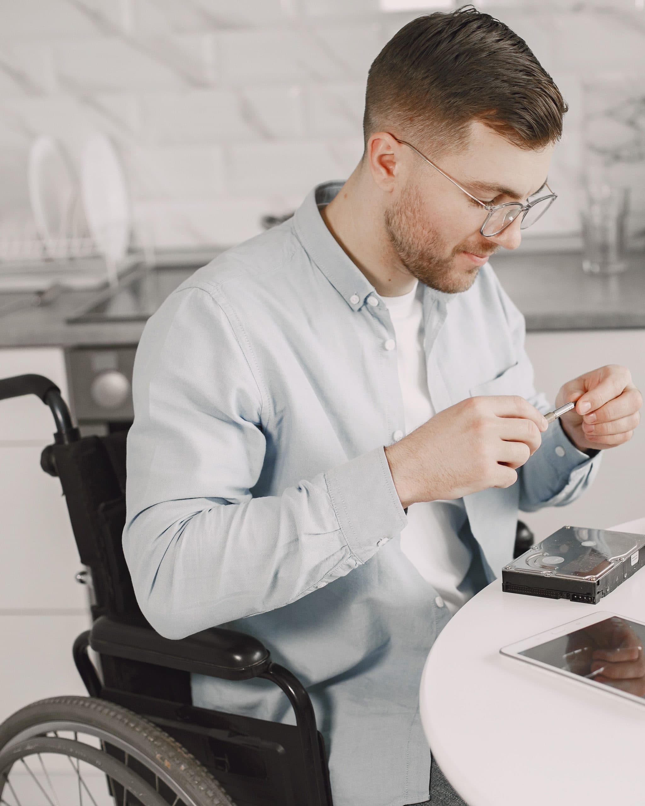 A man in a light blue shirt and glasses sits in a wheelchair at a round white table in a bright kitchen, holding a small part between his fingers beside an opened hard drive and a tablet.