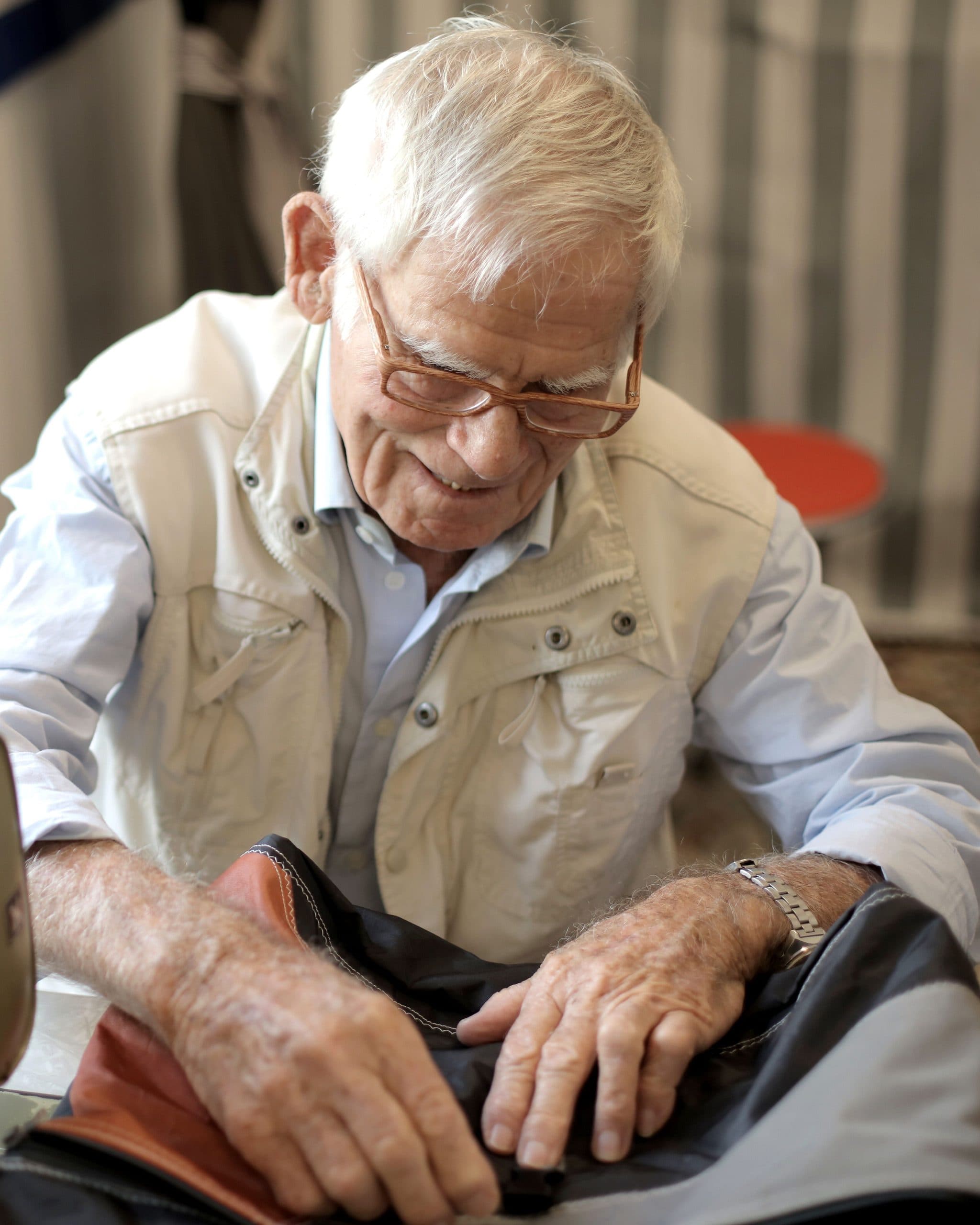 An older man with white hair and glasses smiles as he guides dark fabric under the needle of an industrial sewing machine in a bright workshop.
