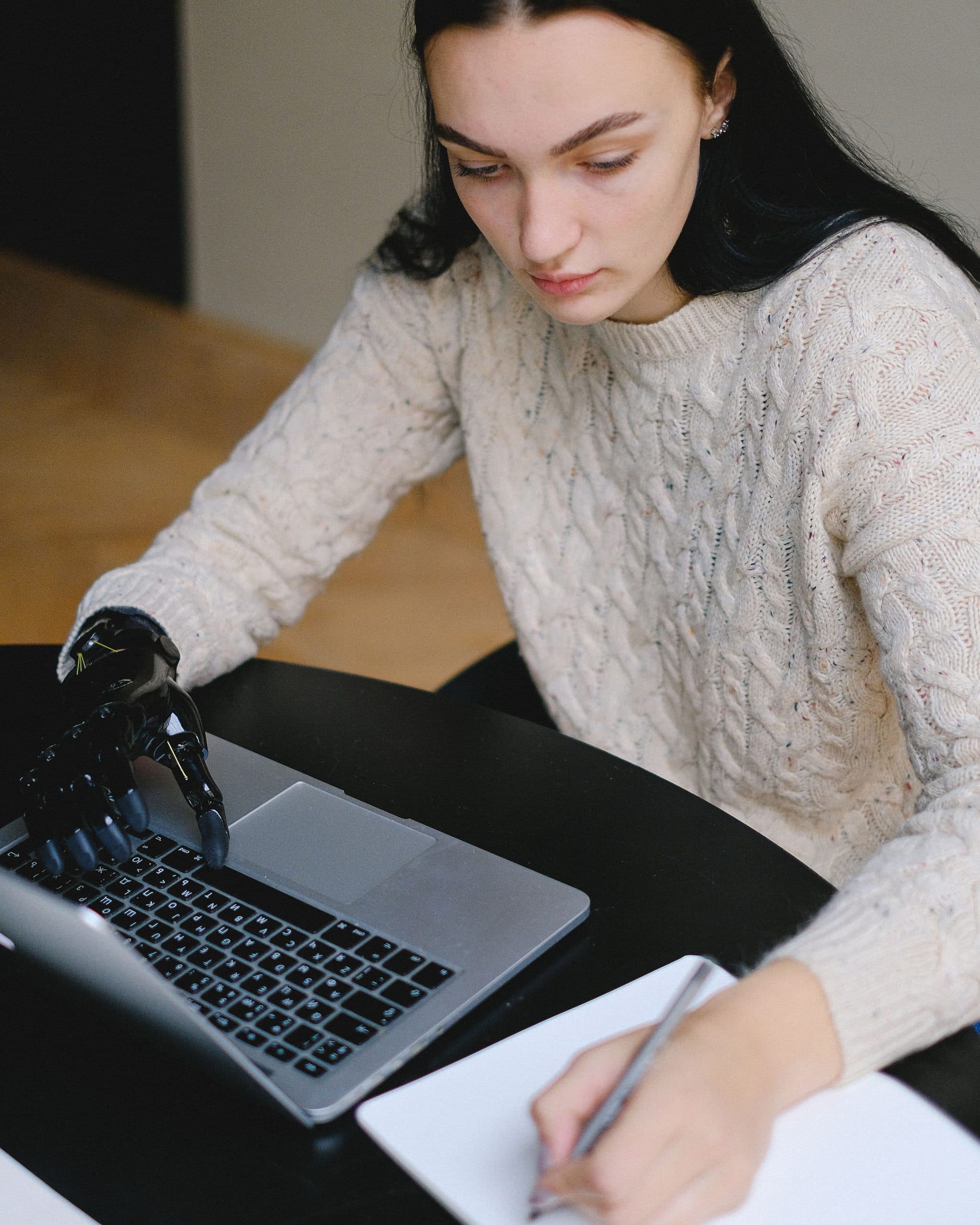 A young woman with long dark hair writes in a notebook beside an open laptop at a dark table. One of her hands is a black prosthetic hand resting on the keyboard.
