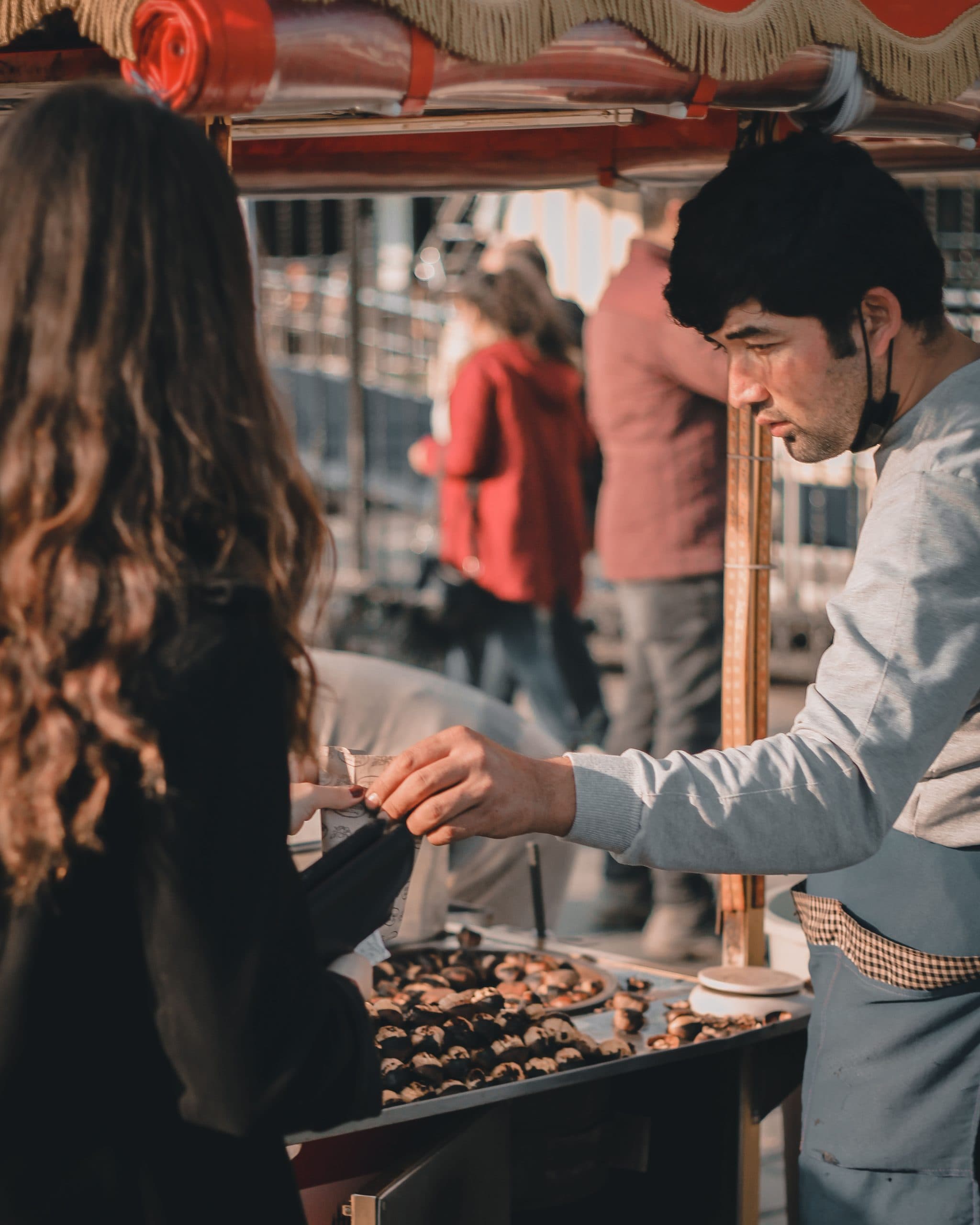 In warm evening light, a street vendor reaches over a stall of roasted chestnuts to serve a customer with long wavy hair, seen from behind.