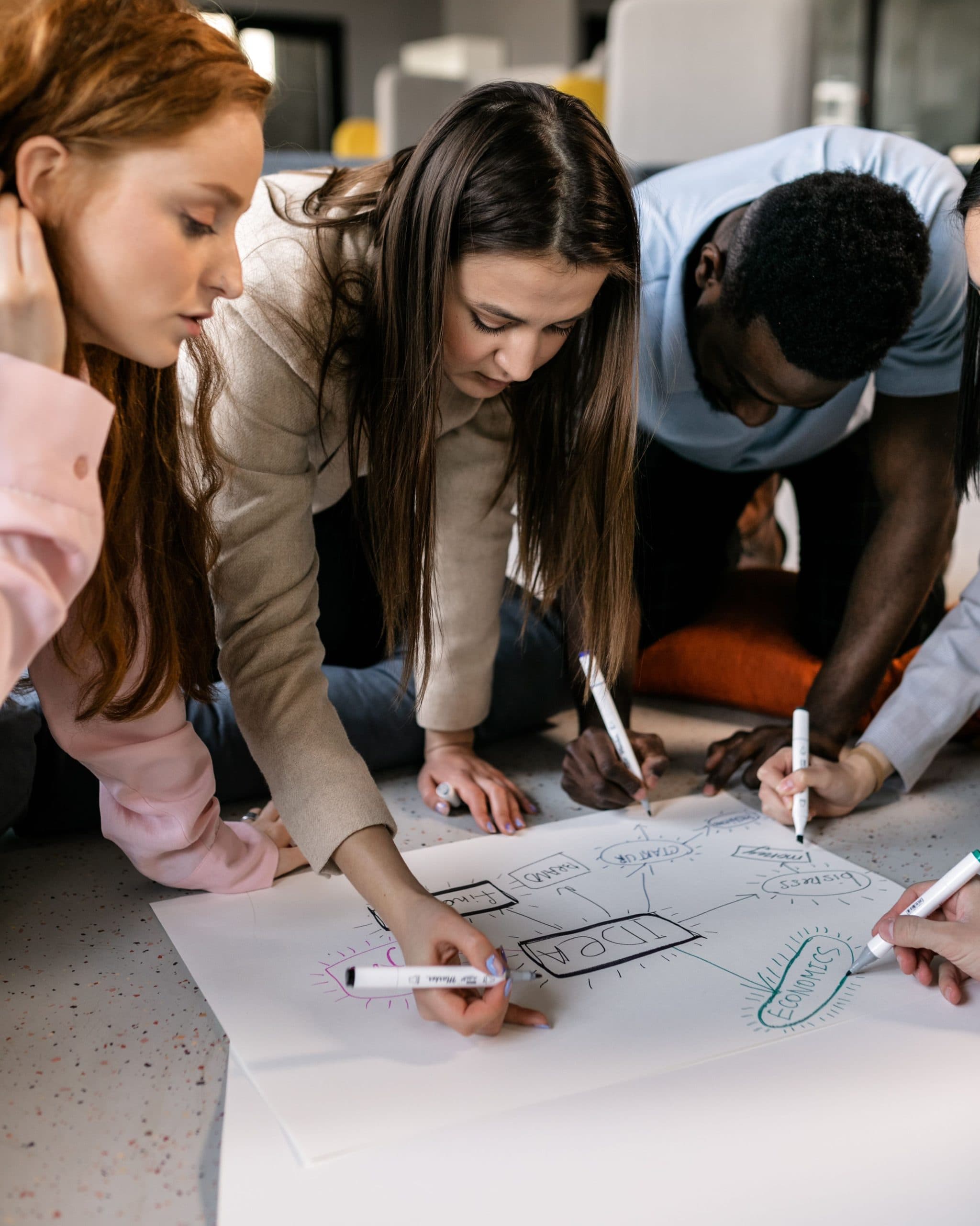A group of people crouch around a large sheet of paper on a pale floor, each drawing on a shared diagram with markers.