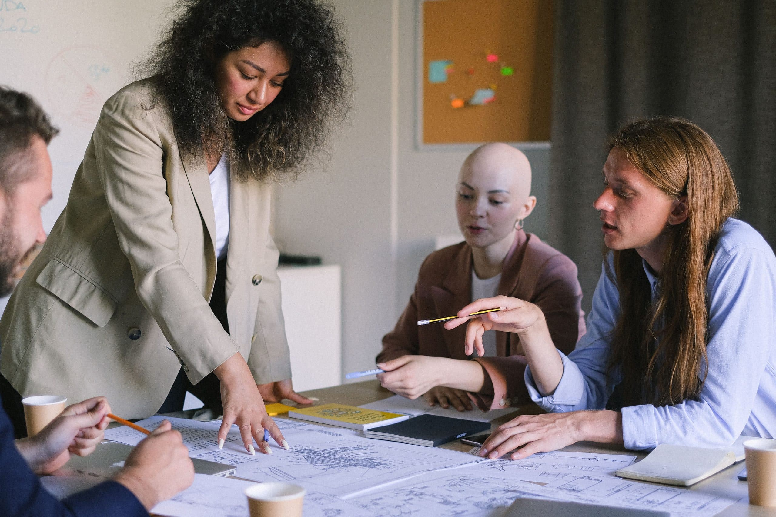 Four people gathered around a table covered in plans and papers, one standing and pointing at the plans while the others lean in to discuss them.