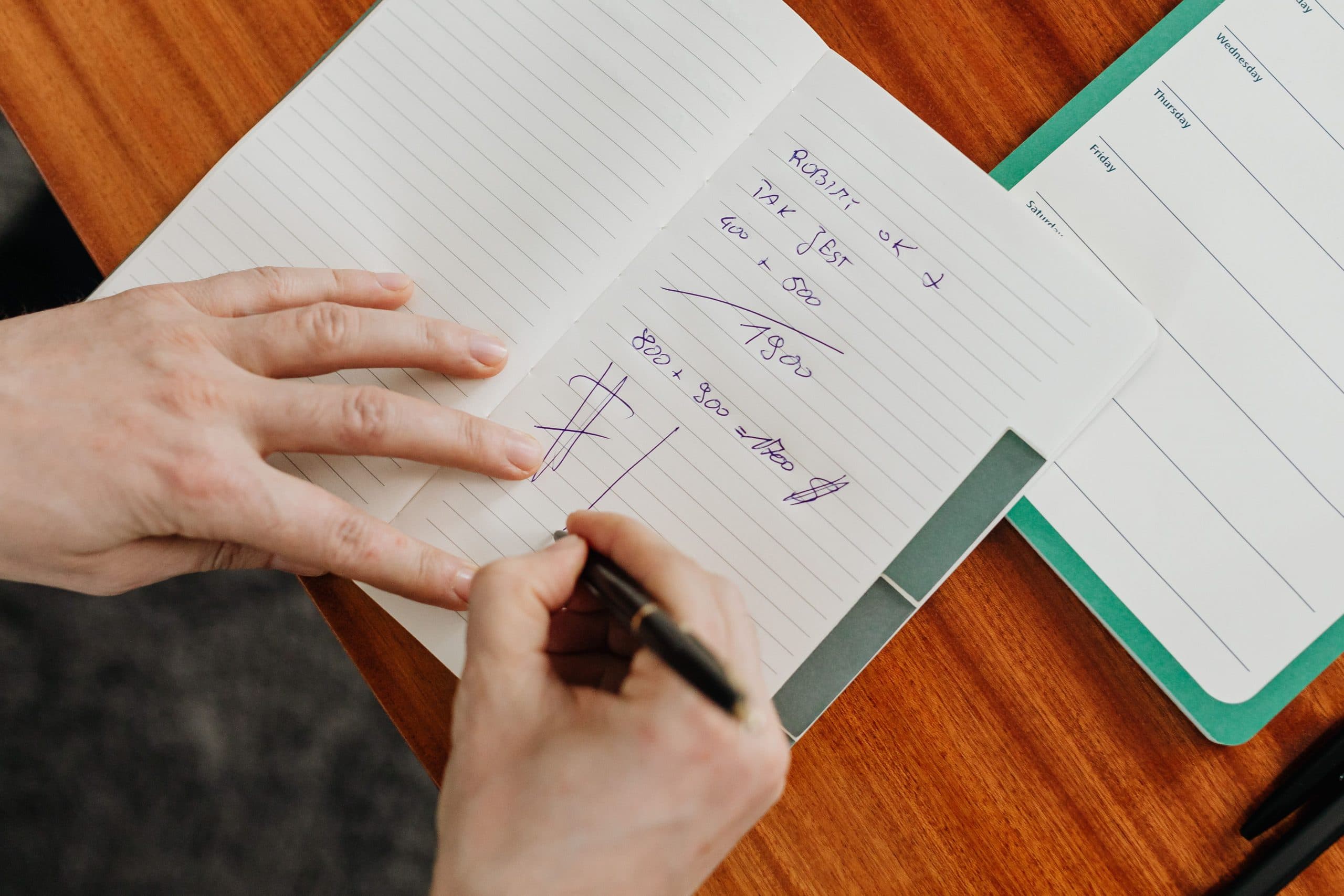 A hand steadies the page of a lined notebook while another hand writes in it, on a warm wooden desk.