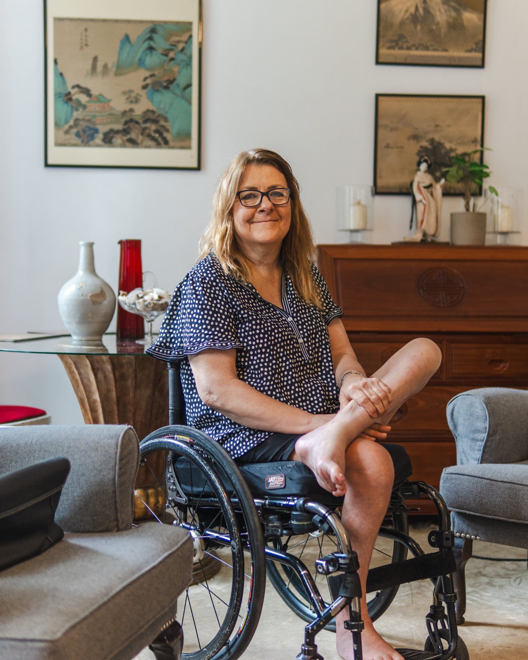 A relaxed woman sits with one leg tucked up in her wheelchair in a bright living room, with framed prints and a wooden cabinet behind her.