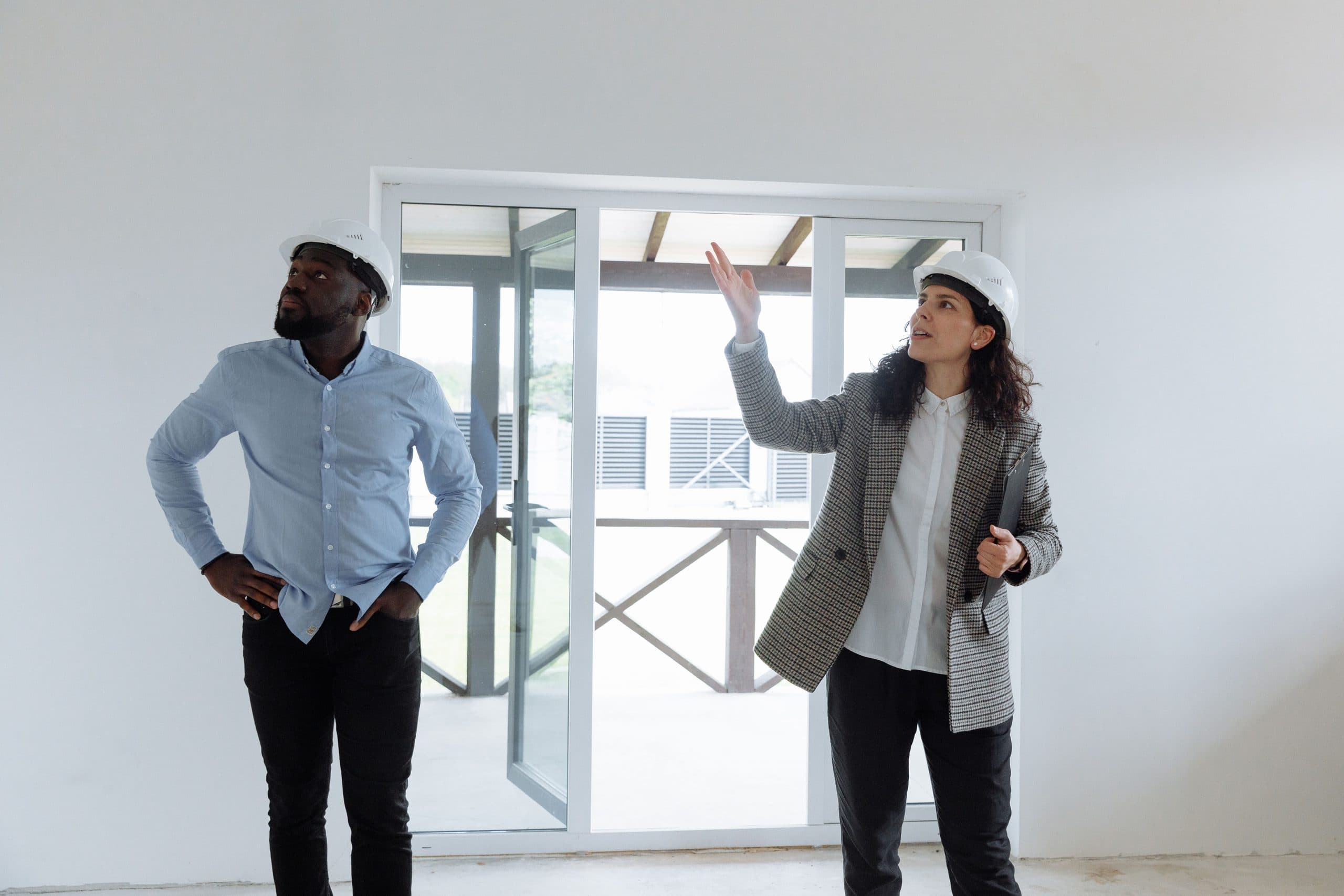 Two people in hard hats stand in a bright, empty, newly finished room with glass doors, one gesturing towards the space.