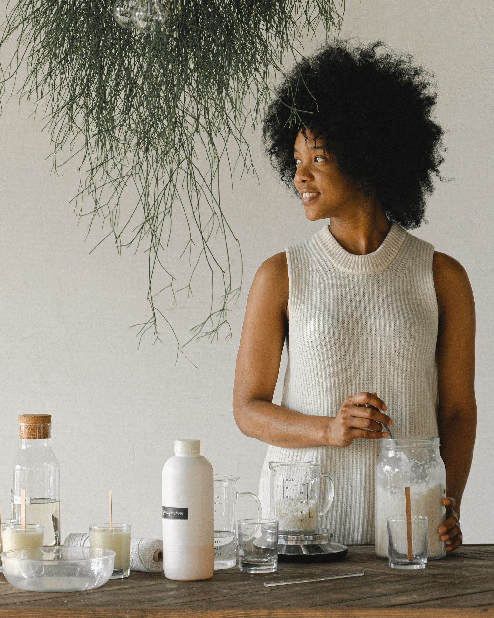 A woman with natural curly hair stands at a long wooden worktable set with jars, glass cups and wax-making equipment in a bright white room, looking off to the side.