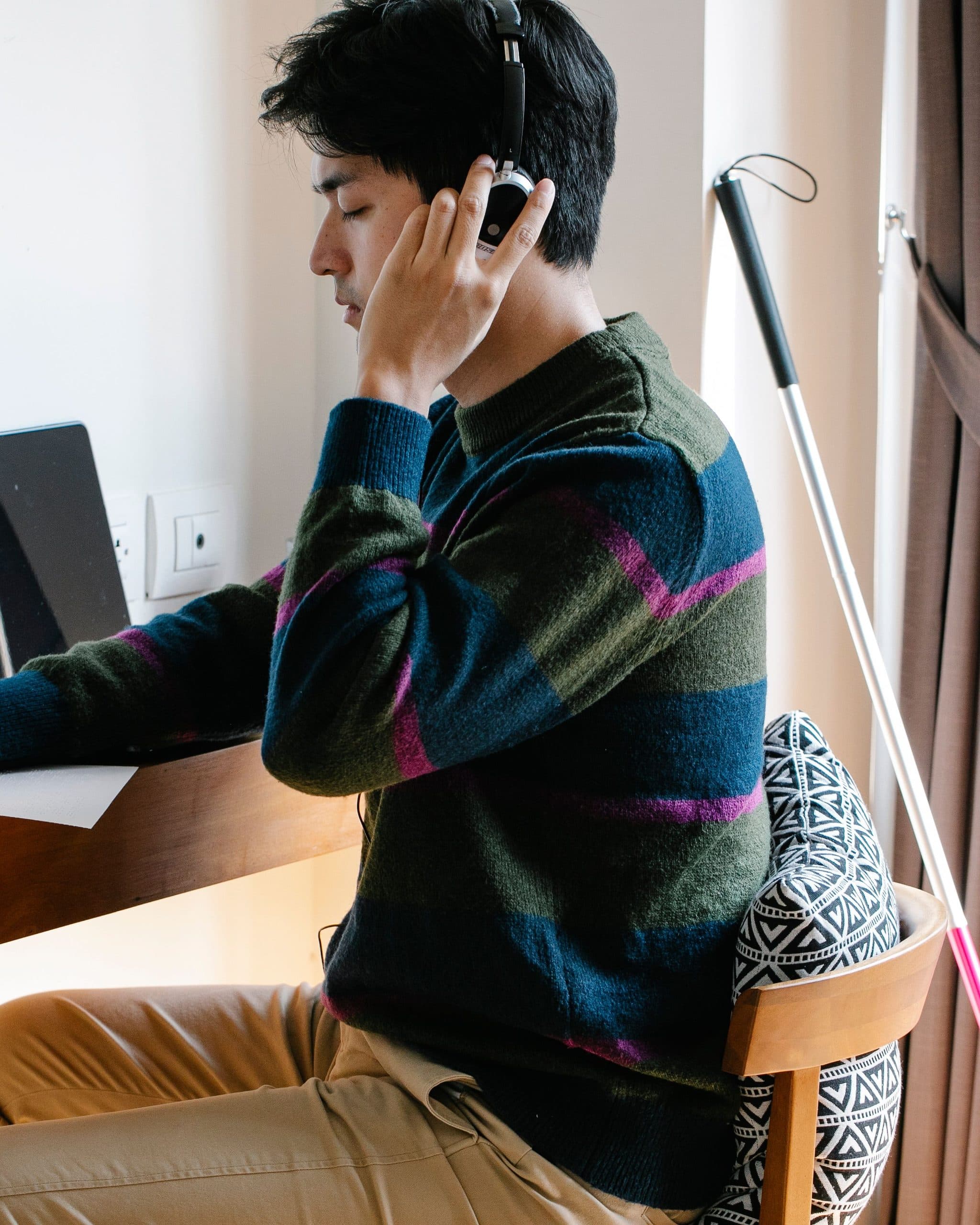 In profile, a man wearing over-ear headphones rests one hand on a sheet of paper at a wall-mounted desk beside a window. A white cane hangs by the curtain.
