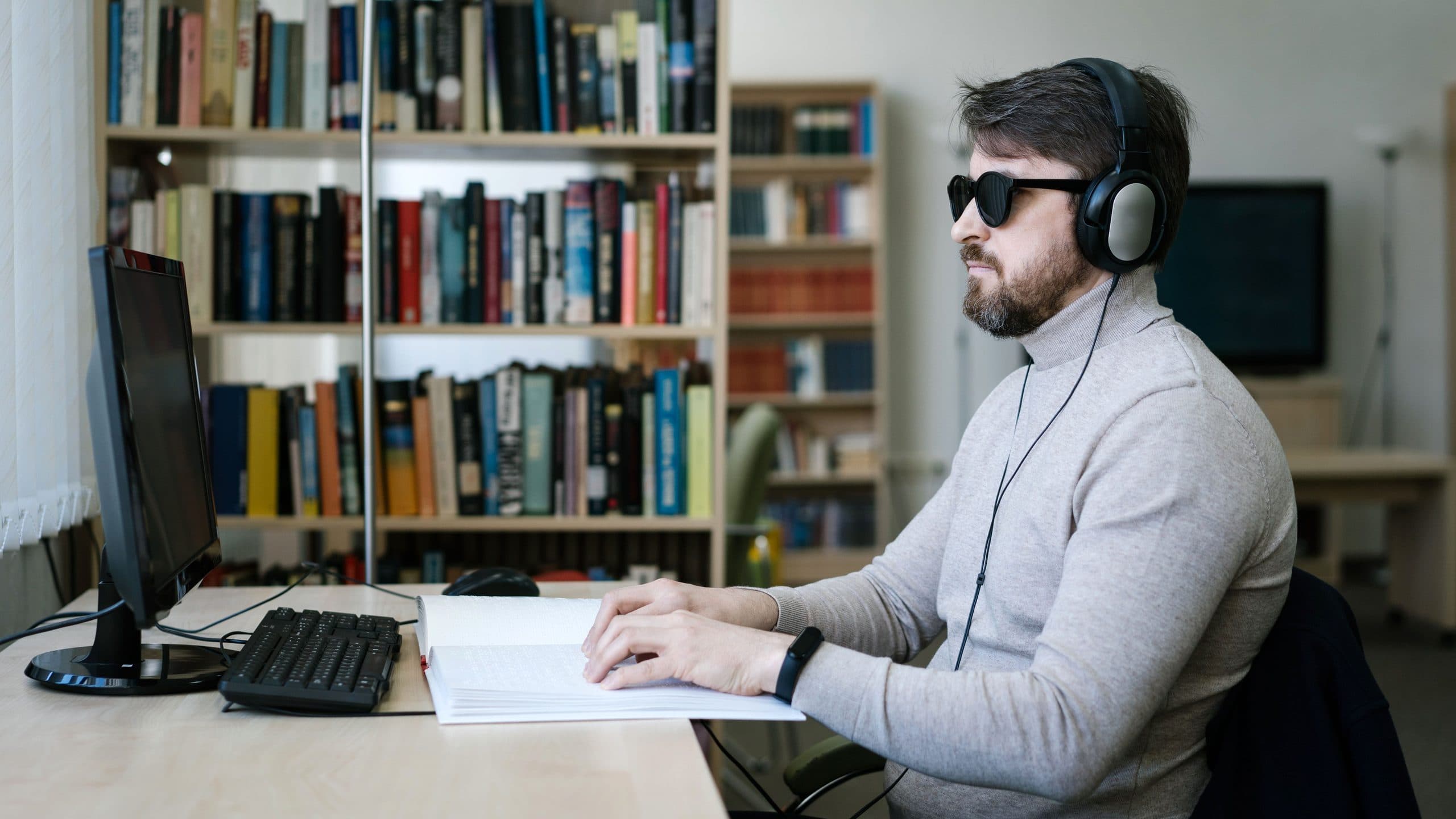 A man wearing dark glasses and over-ear headphones sits at a desk in a library, one hand on an open braille book and the other near a computer keyboard.