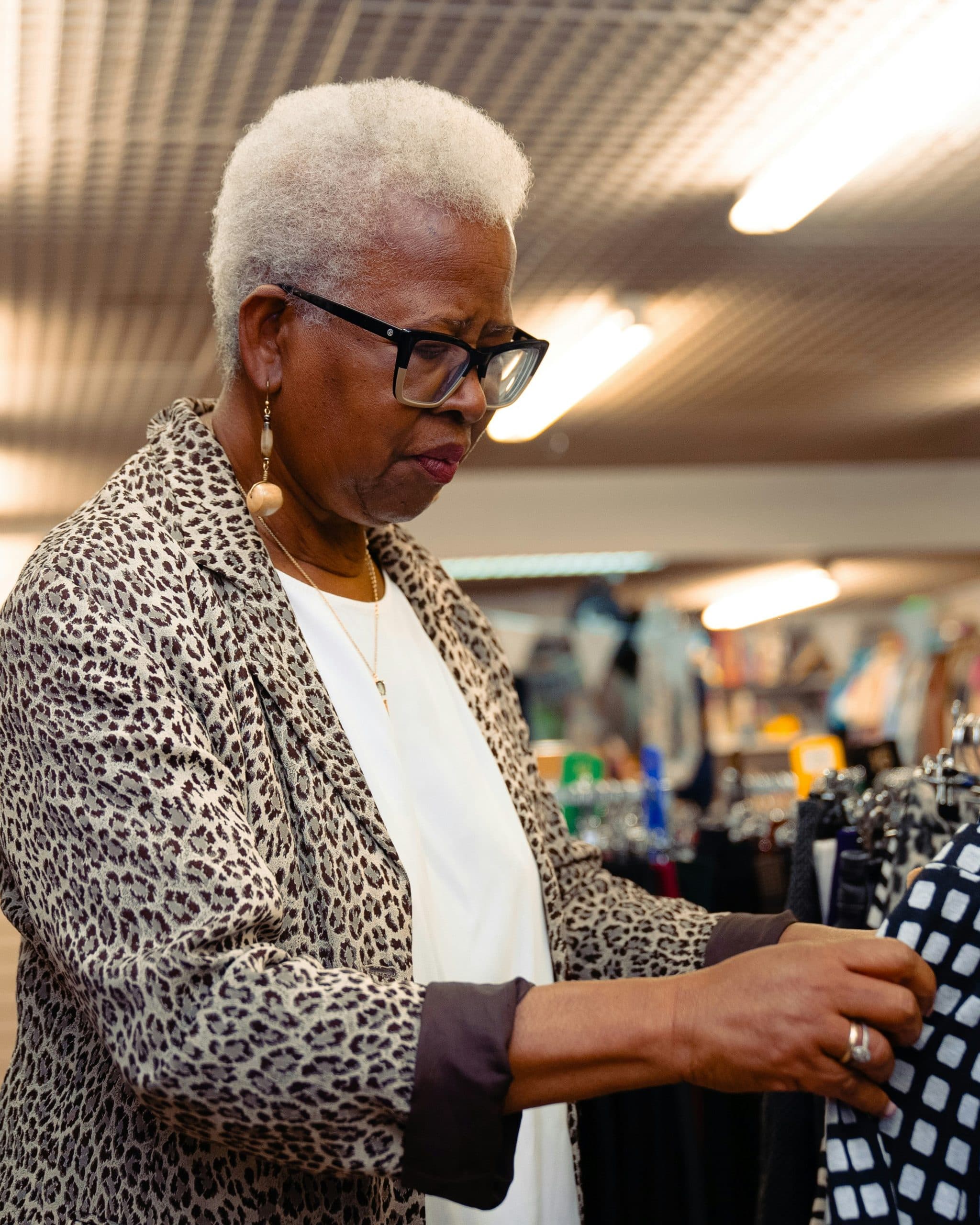 An older woman with short white hair and glasses, in a leopard-print jacket, looks through garments on a clothes rail in a shop.