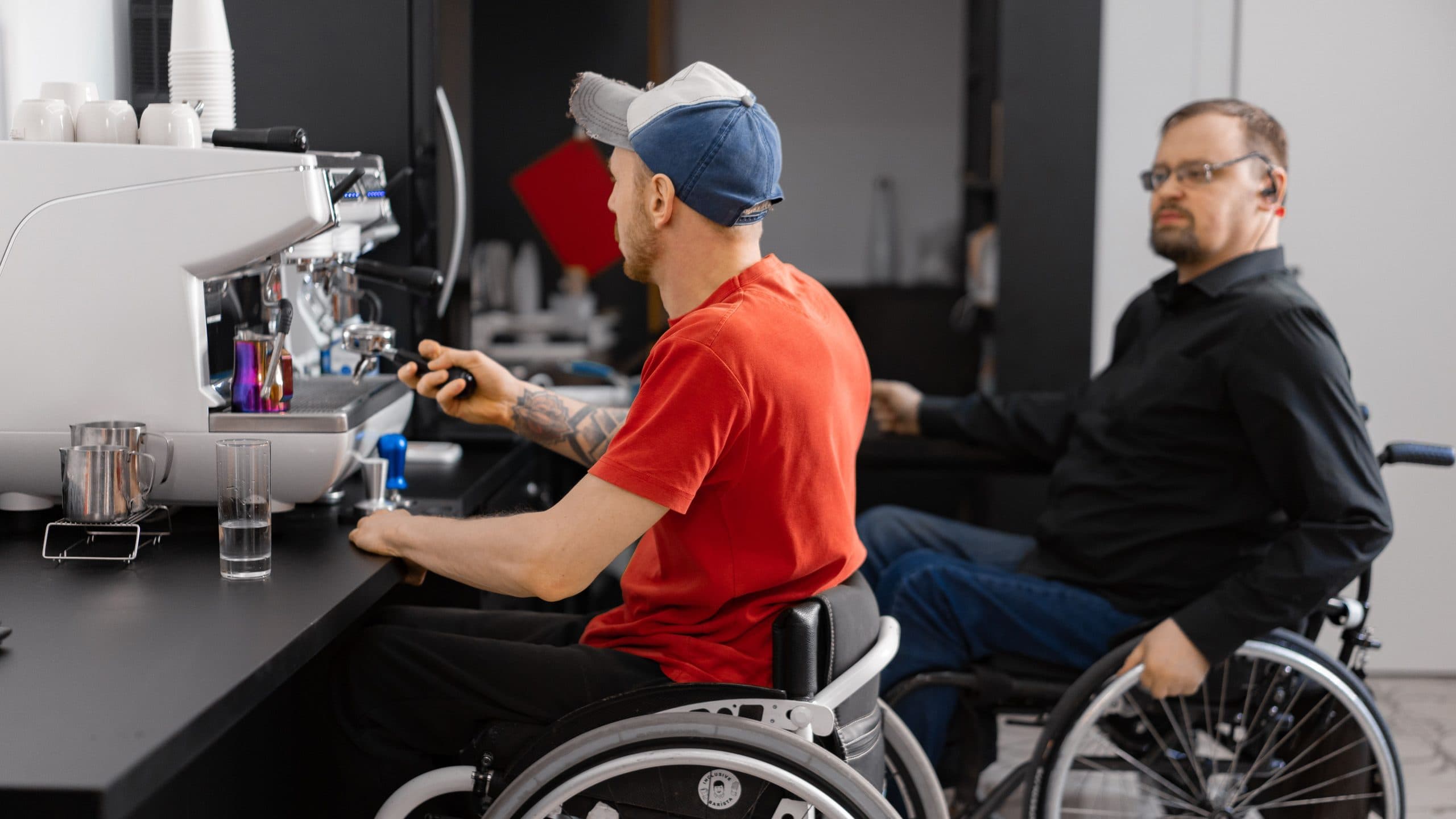 Two men seated in wheelchairs work side by side at the counter of a cafe. One, in a red T-shirt and a cap, works the controls of an espresso machine; the other, in a black shirt and glasses, sits back from the counter and looks toward the camera.