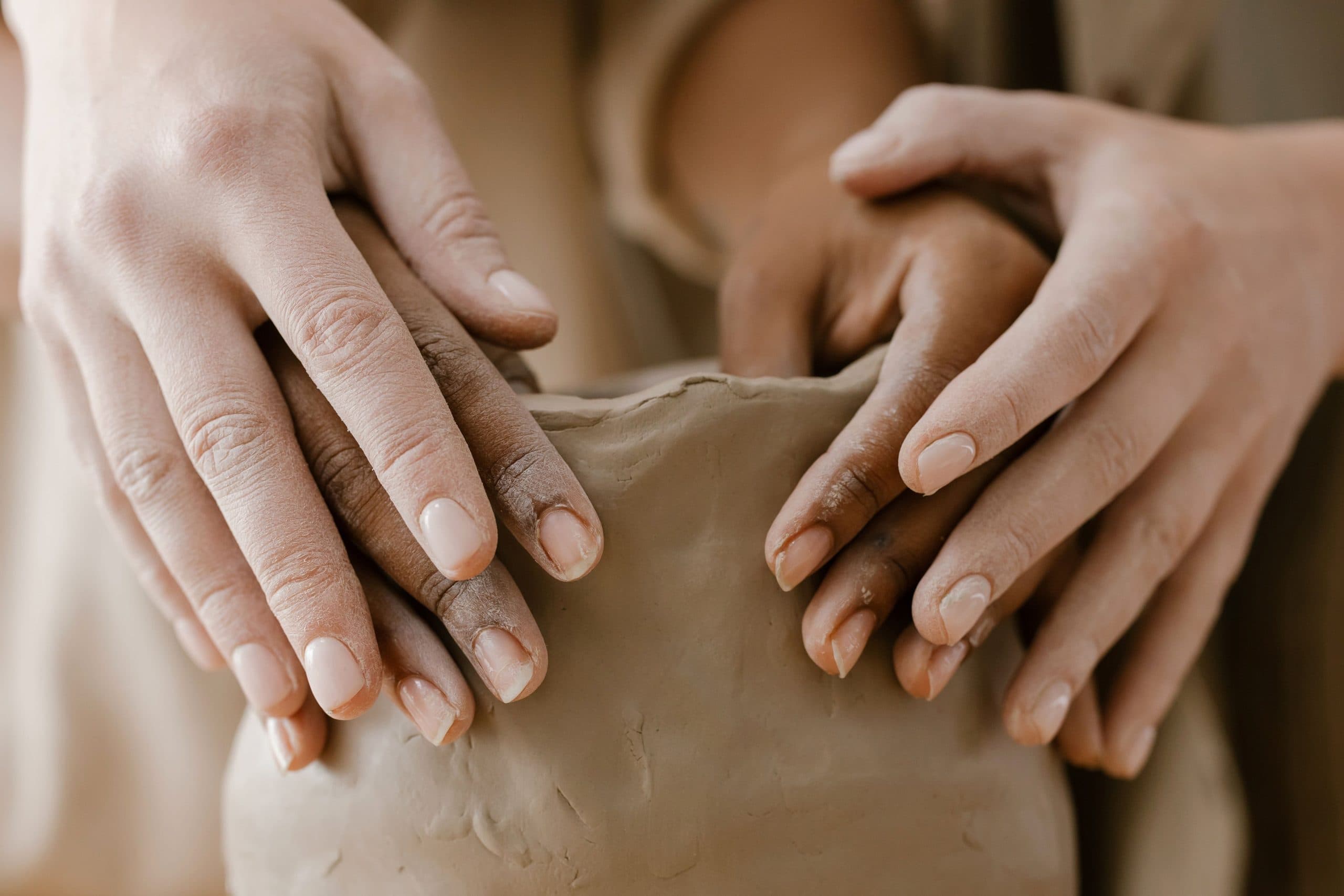 Close view of several hands of different skin tones shaping a clay pot together.