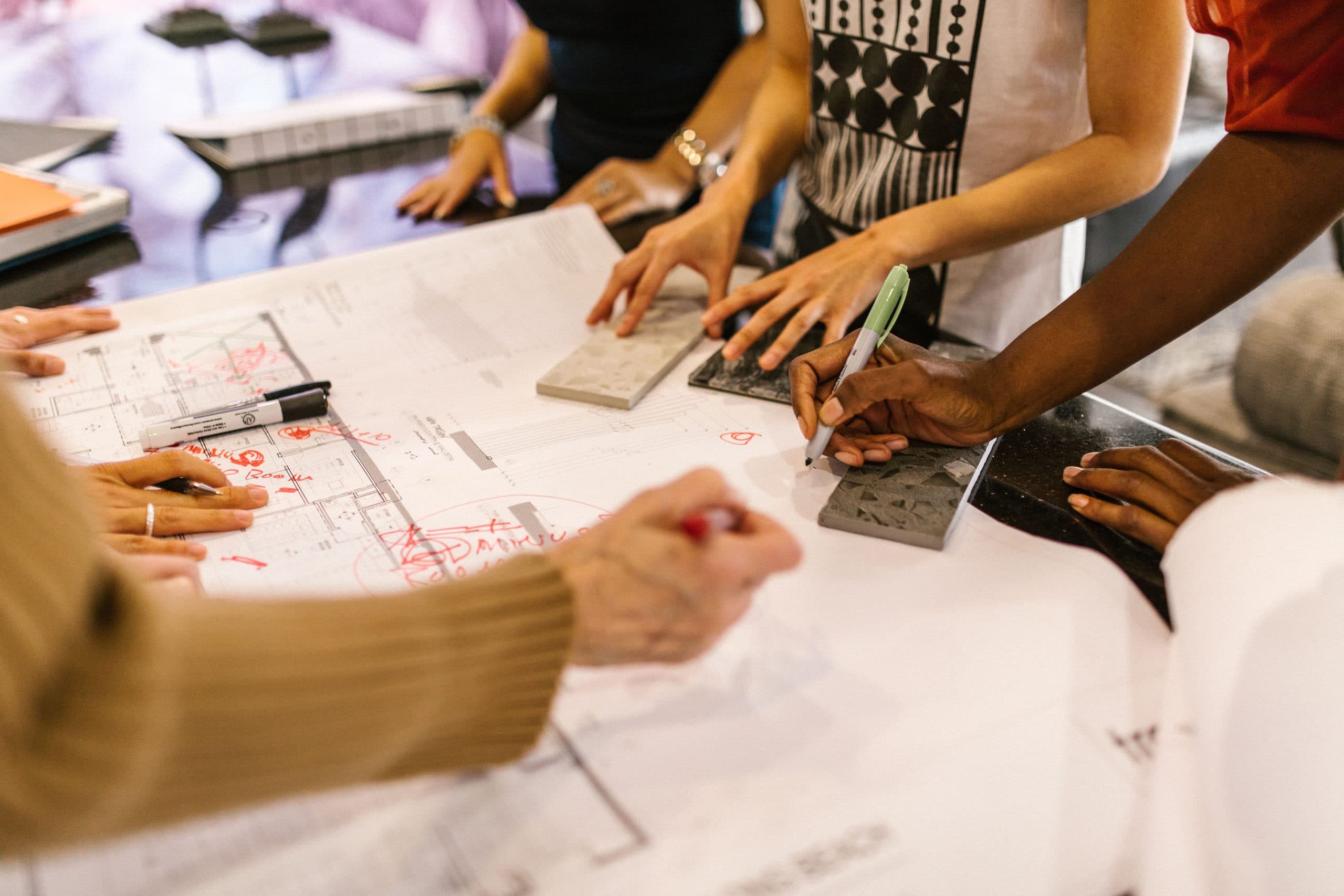 Several pairs of hands rest on a large floor plan laid on a table, marking it with a red pen and placing tile samples on the drawing.