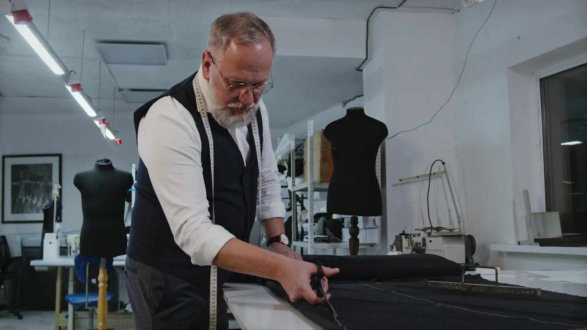 A man with a grey beard and glasses, wearing a waistcoat with the sleeves of his white shirt rolled up, cuts black cloth with large shears along a long cutting table in a bright tailoring workshop. Dress forms and sewing machines stand behind him.