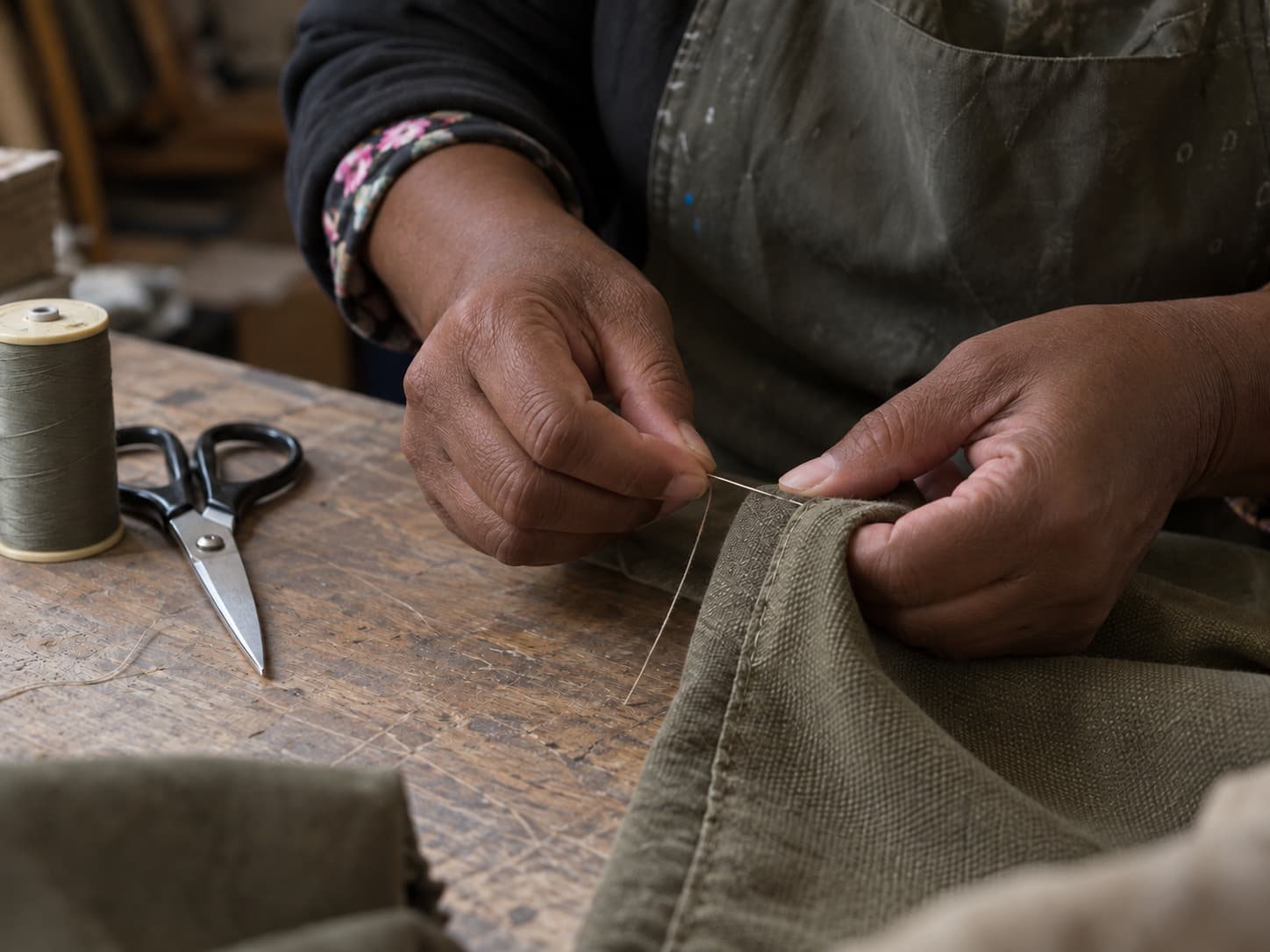 Close view of hands stitching heavy olive fabric by hand, with thread and scissors on the bench.