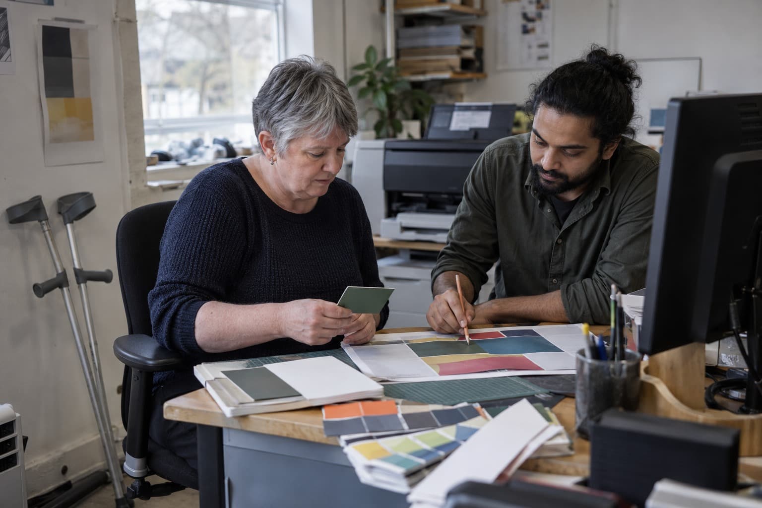 Two colleagues compare printed colour swatches at a studio desk, crutches resting beside the chair.