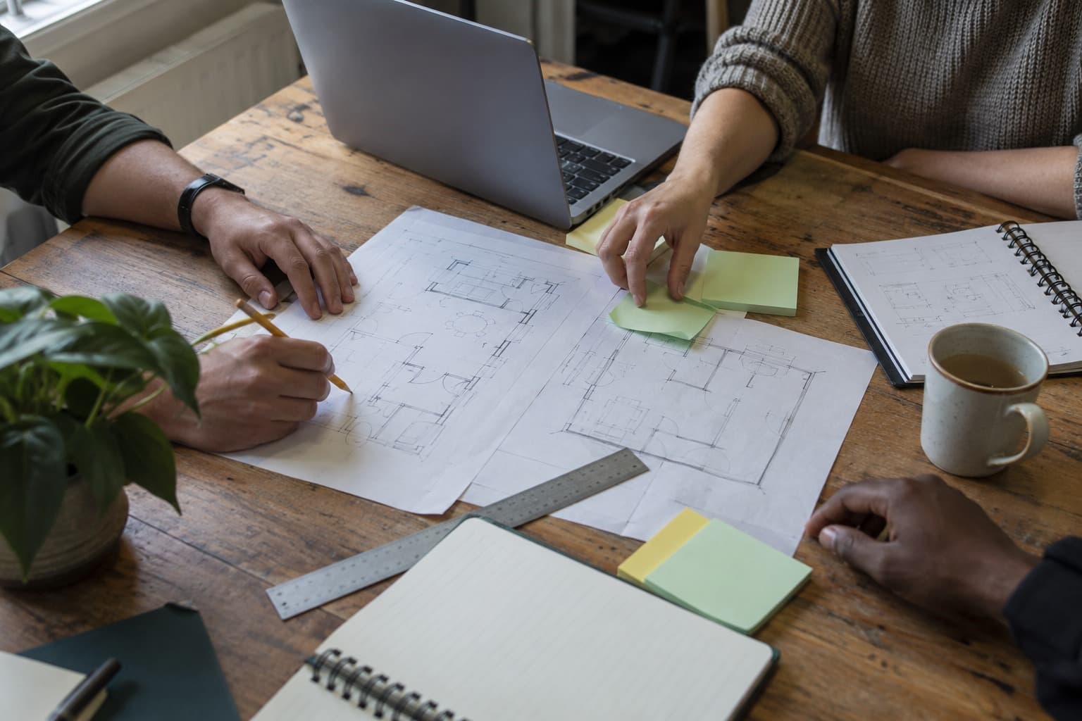 Hands annotating architectural floor plans beside a laptop, notebooks and sticky notes.